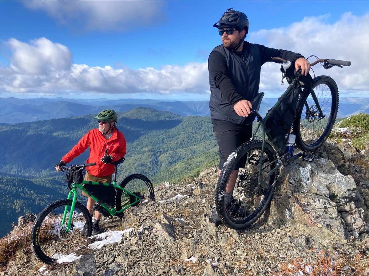 bikerumor pic of the day tow mountain bikers on a peak overlooking a lush valley below.