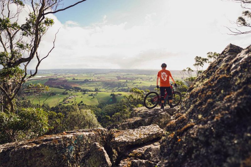 un mountain biker in piedi con la sua bici in cima a un affioramento roccioso che domina una vasta valle verde